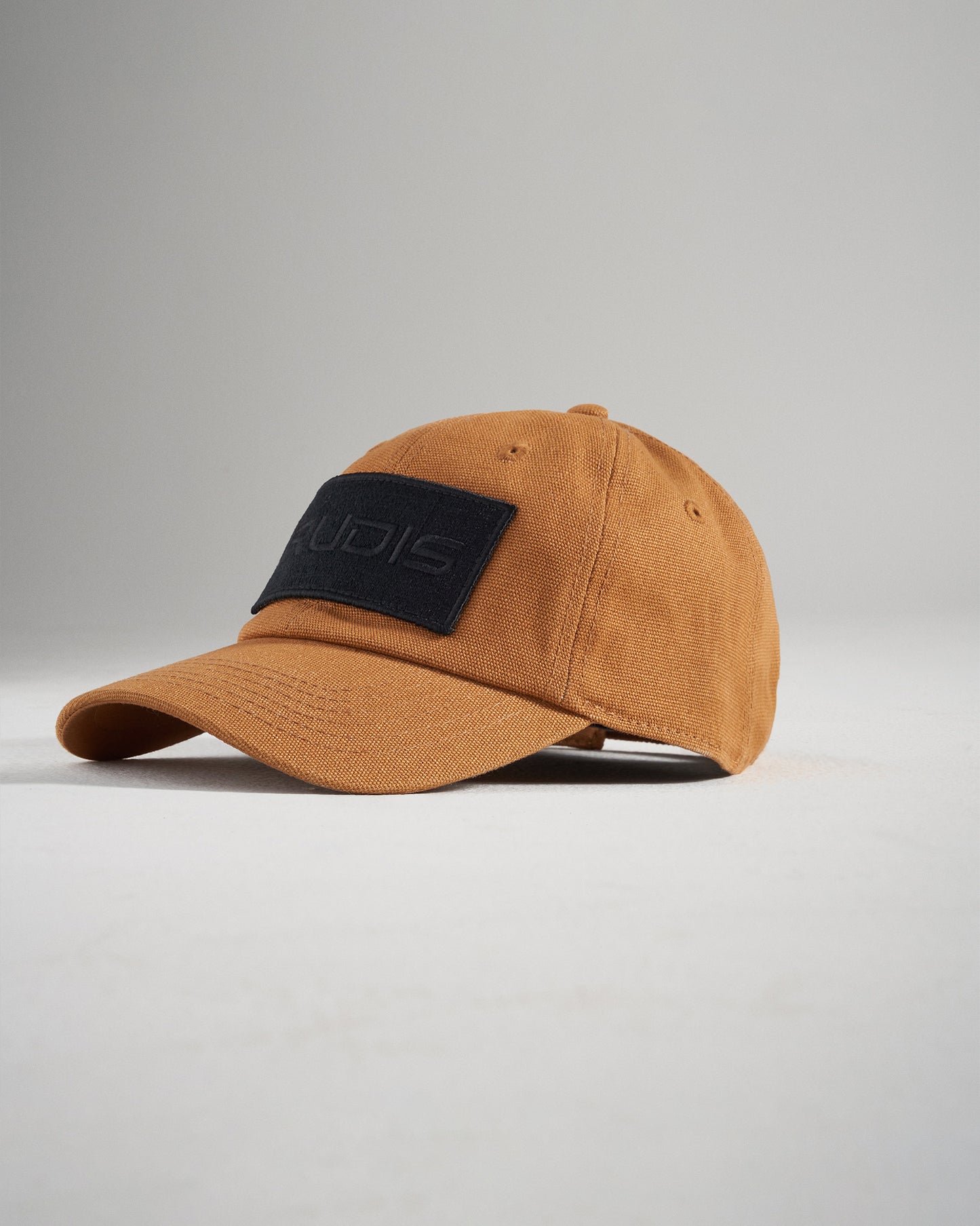 A stylish brown cap featuring a black patch with the word RUDIS in bold lettering set against a minimalist background