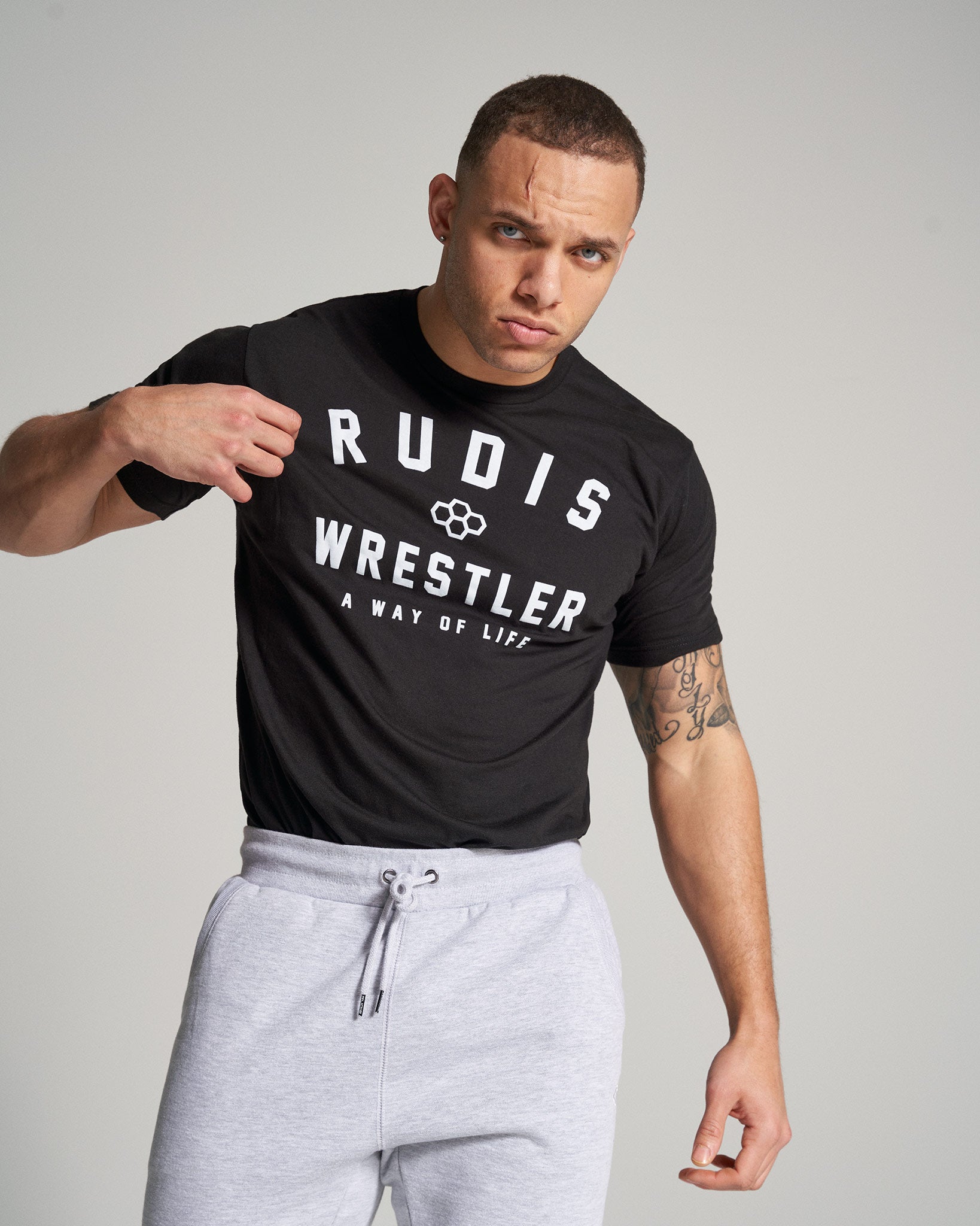 A fit man wearing a black t-shirt with RUDIS WRESTLER A WAY OF LIFE text and gray sweatpants showcasing confidence with a serious expression