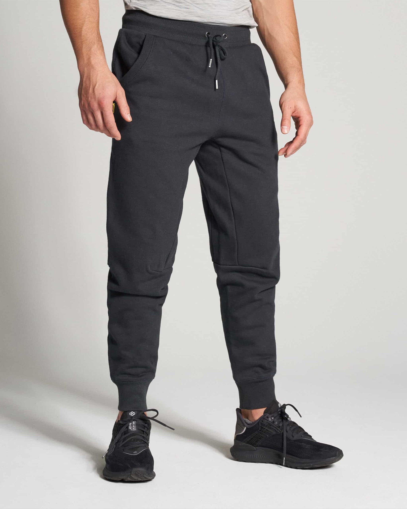 A man in athletic black sweatpants and black sneakers stands against a neutral background showcasing a casual yet sporty look