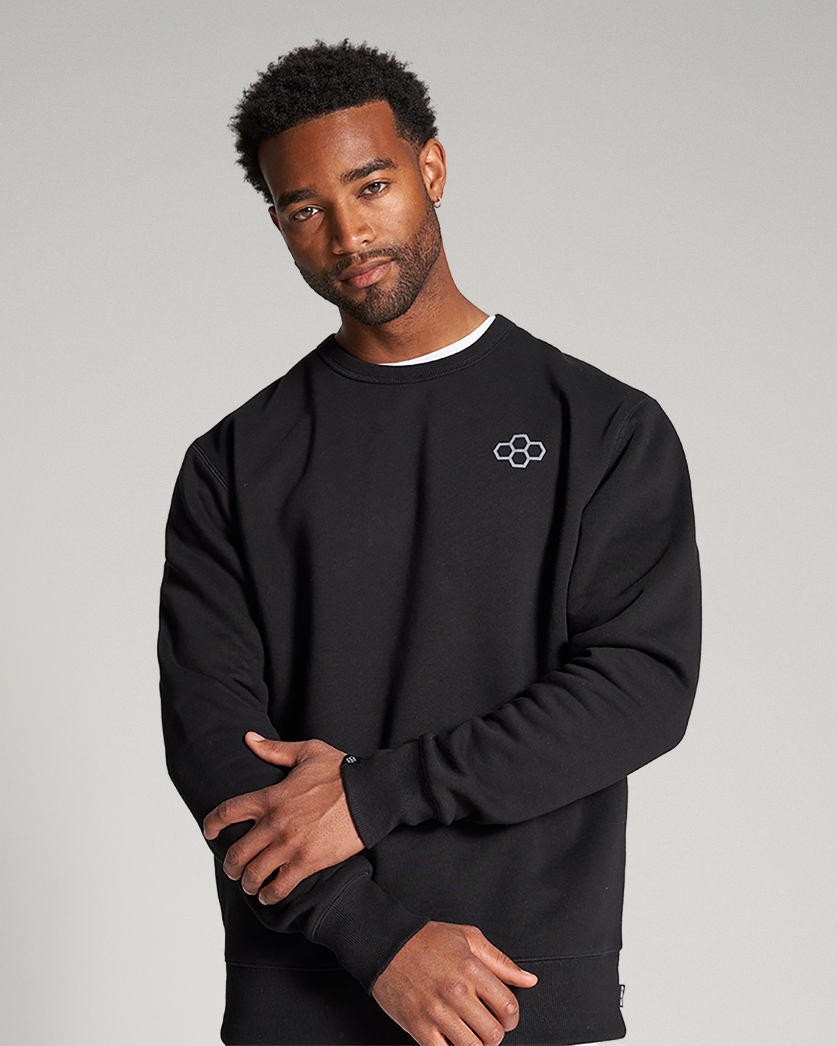 A portrait of a man wearing a black sweatshirt with a round neckline showcasing a relaxed fit and a minimalist flower logo on the chest