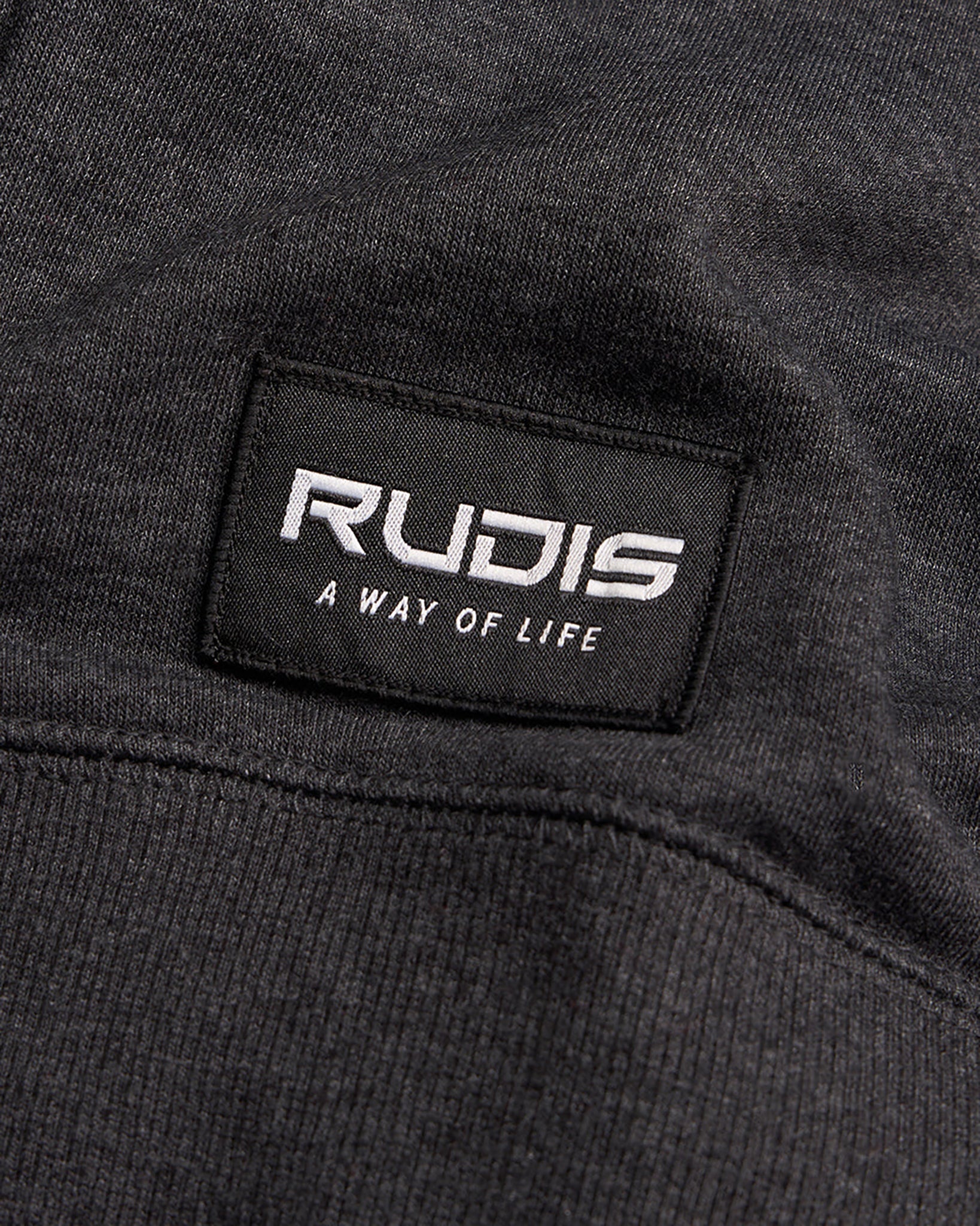 A close-up view of a black fabric label stitched onto a garment featuring the word RUDIS and the phrase A WAY OF LIFE in white text