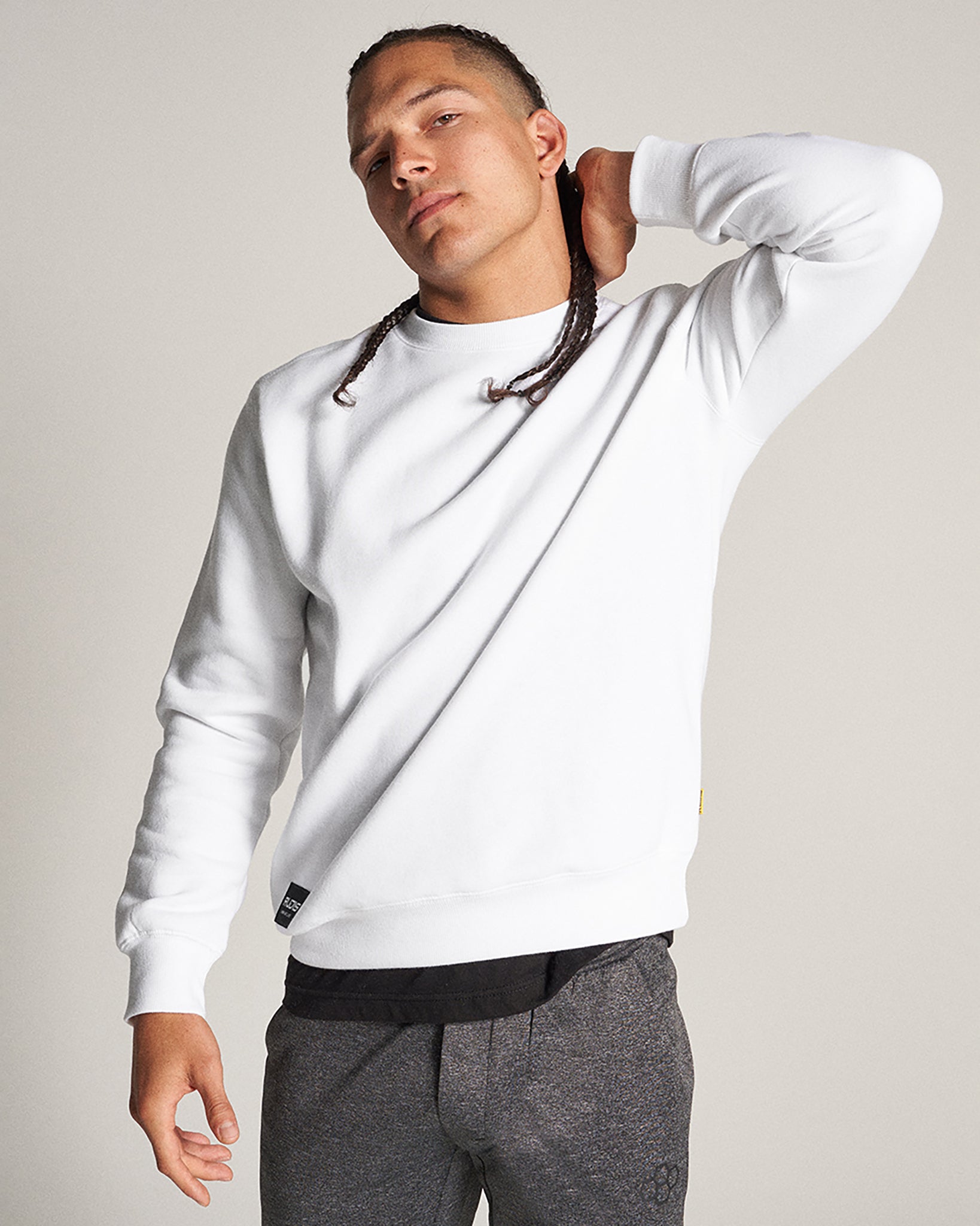 A man wearing a white sweatshirt and gray joggers confidently poses with one hand behind his neck presenting a stylish casual look