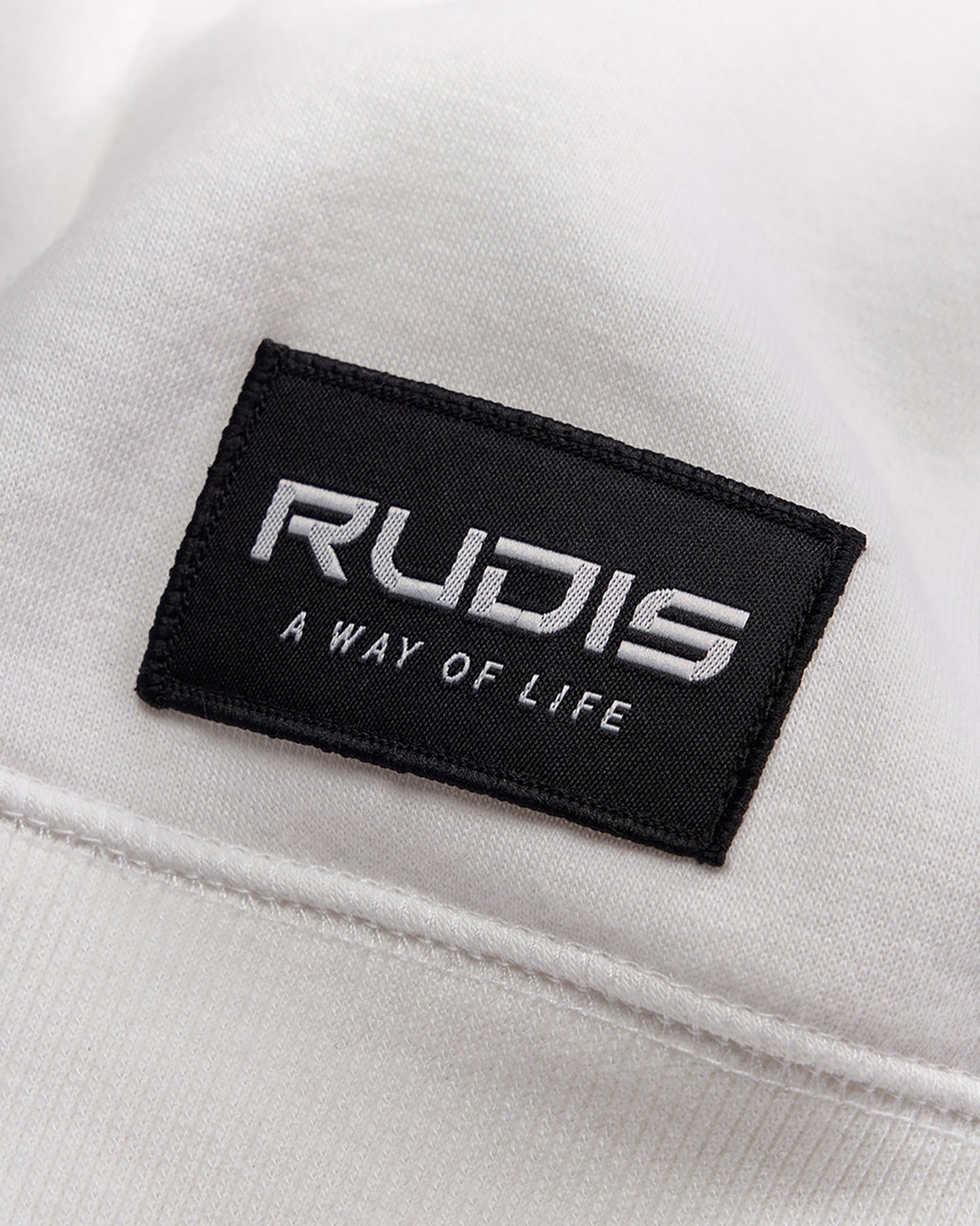 A close-up of a white fabric featuring a black rectangular tag that reads RUDIS A WAY OF LIFE in bold lettering