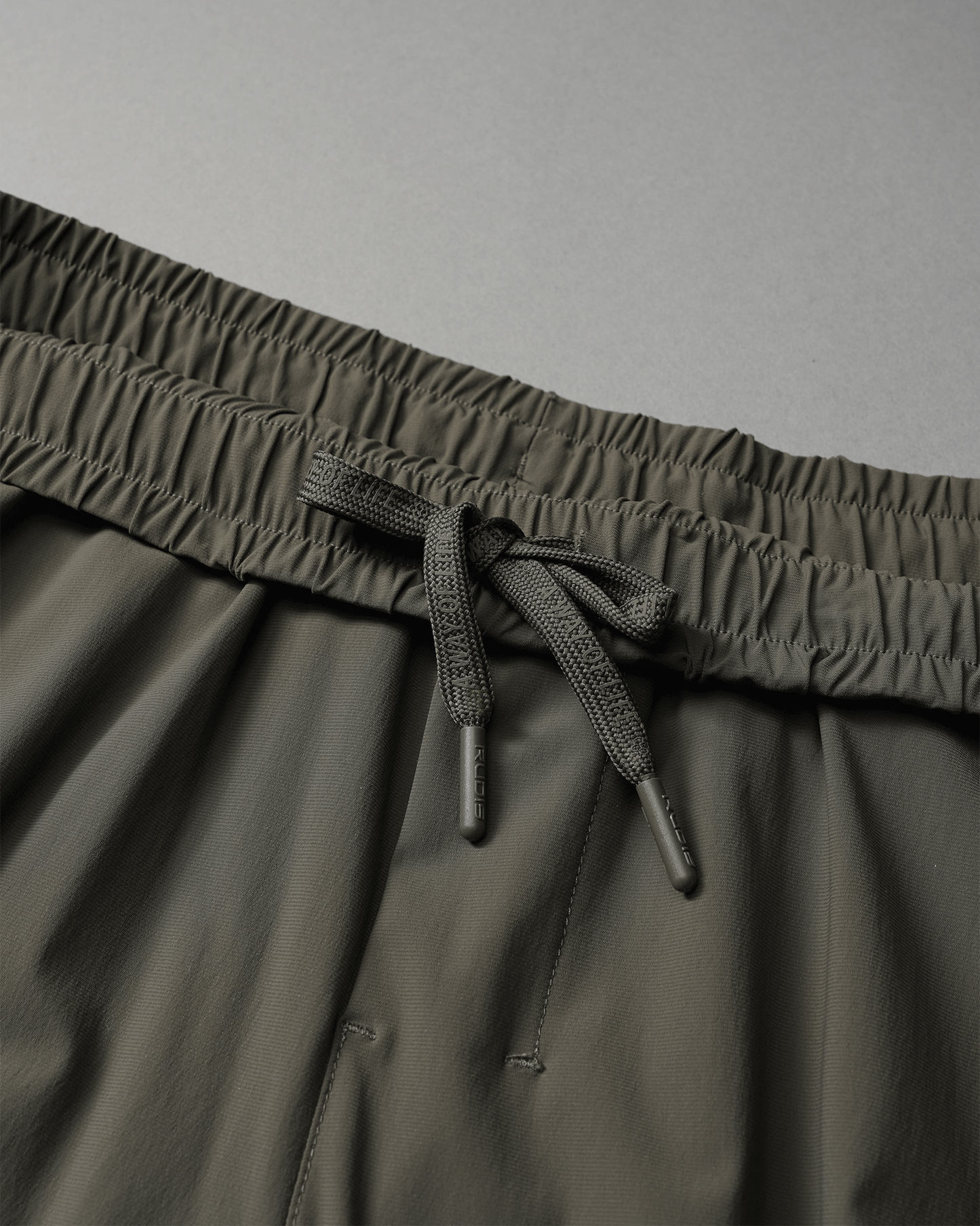 Close-up of dark olive shorts' waistband showing an elastic band and textured drawstring for adjustment against a soft gray background.