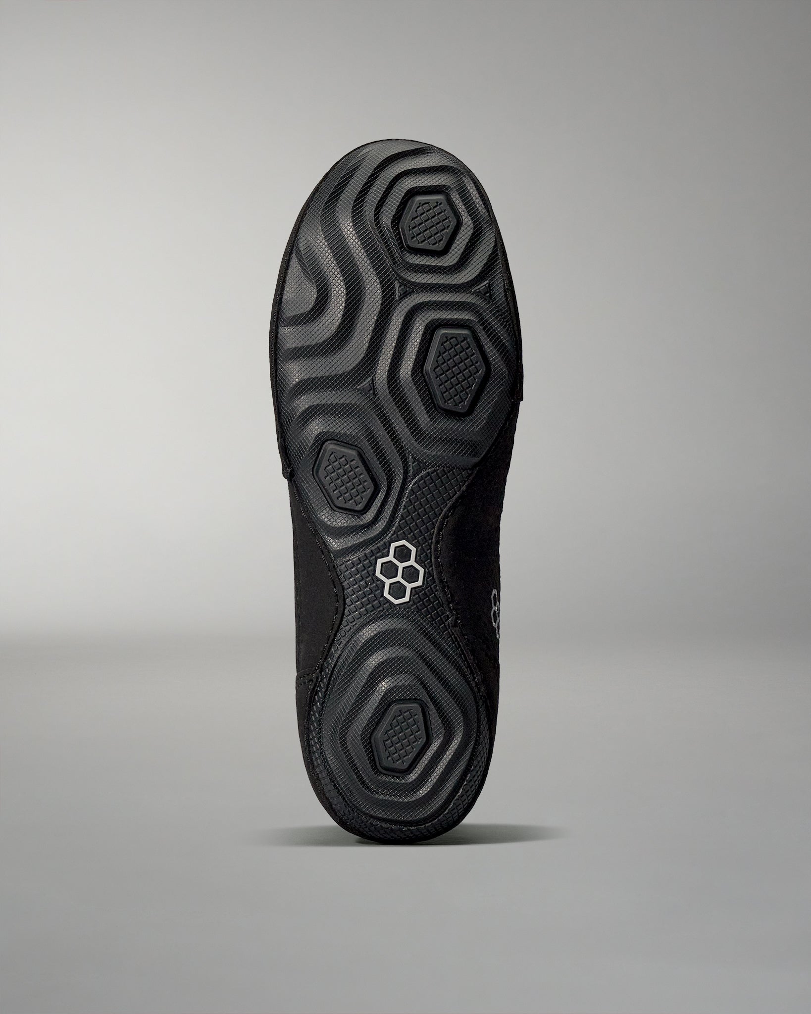 The bottom of a black shoe features a textured sole with hexagonal and circular patterns for enhanced grip, set against a soft gray background.