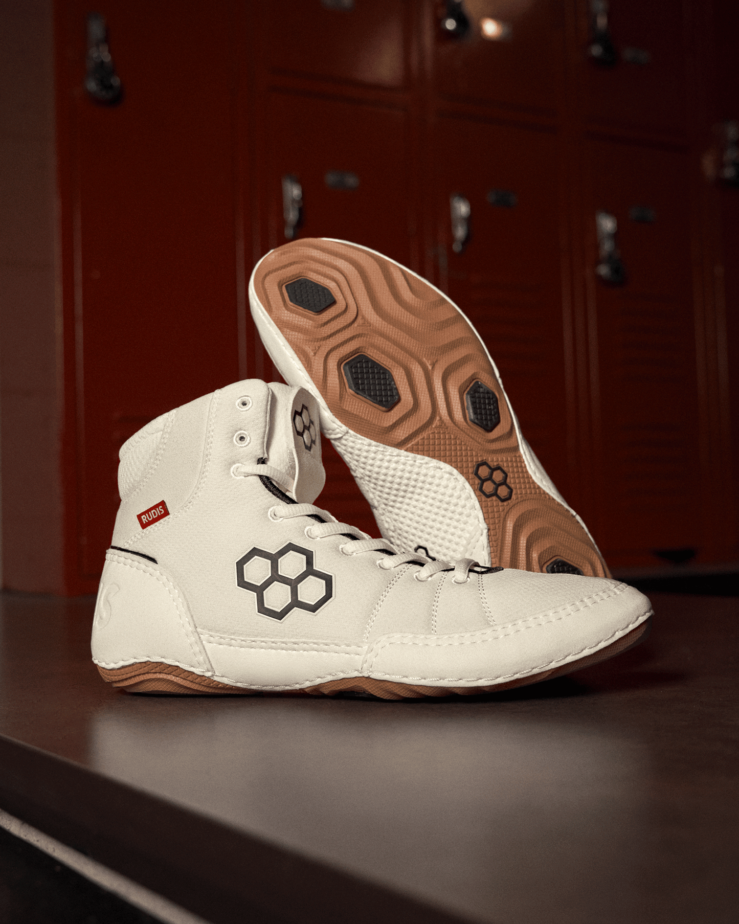 White athletic boots featuring a hexagonal logo and a red 'RUDIS' tag are showcased on a dark surface, with red lockers in the background enhancing their sporty look.