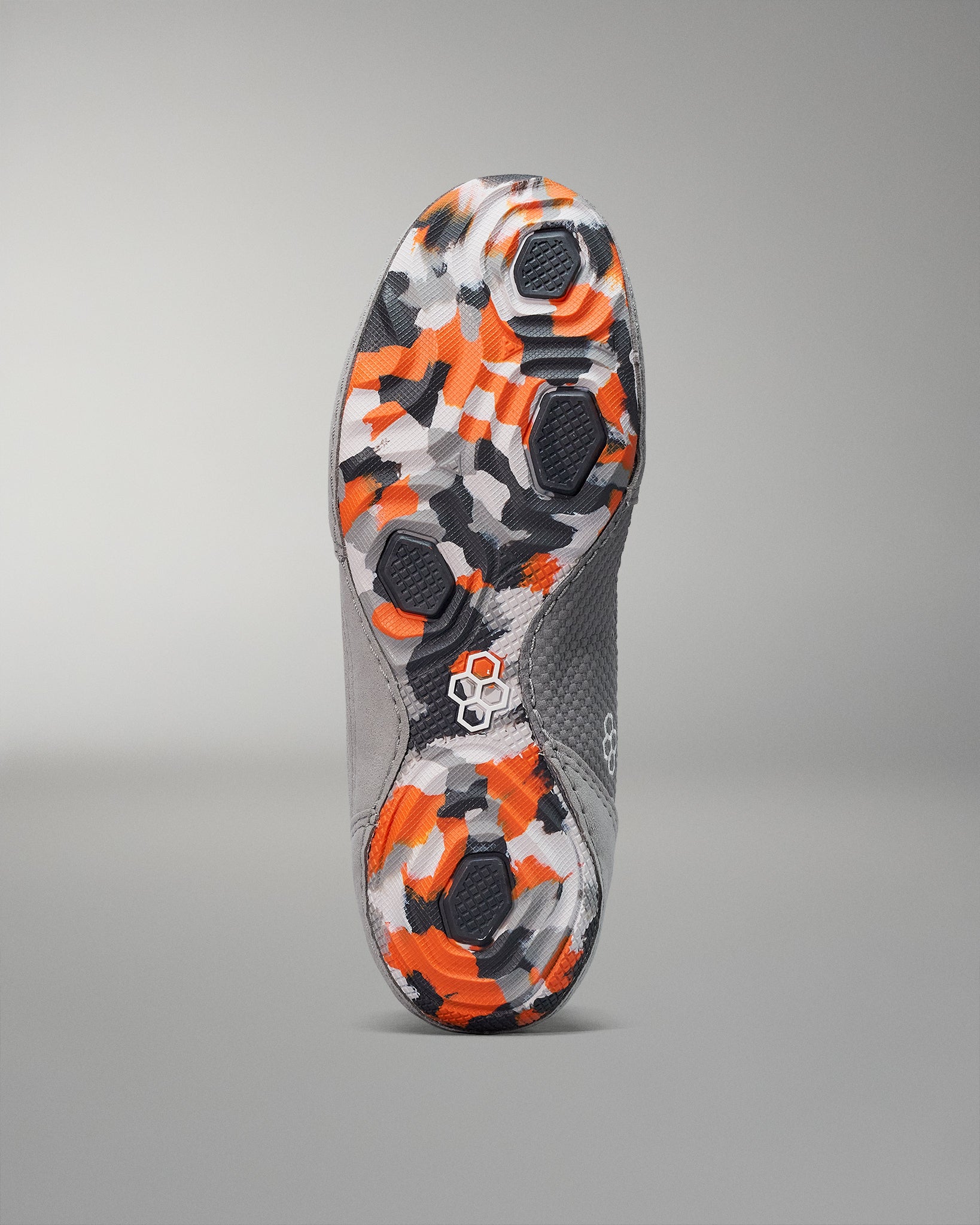 The sole of a shoe features a vibrant camouflage design in orange, black, and gray, with textured grip elements for enhanced traction against a muted gray upper and subtle background.