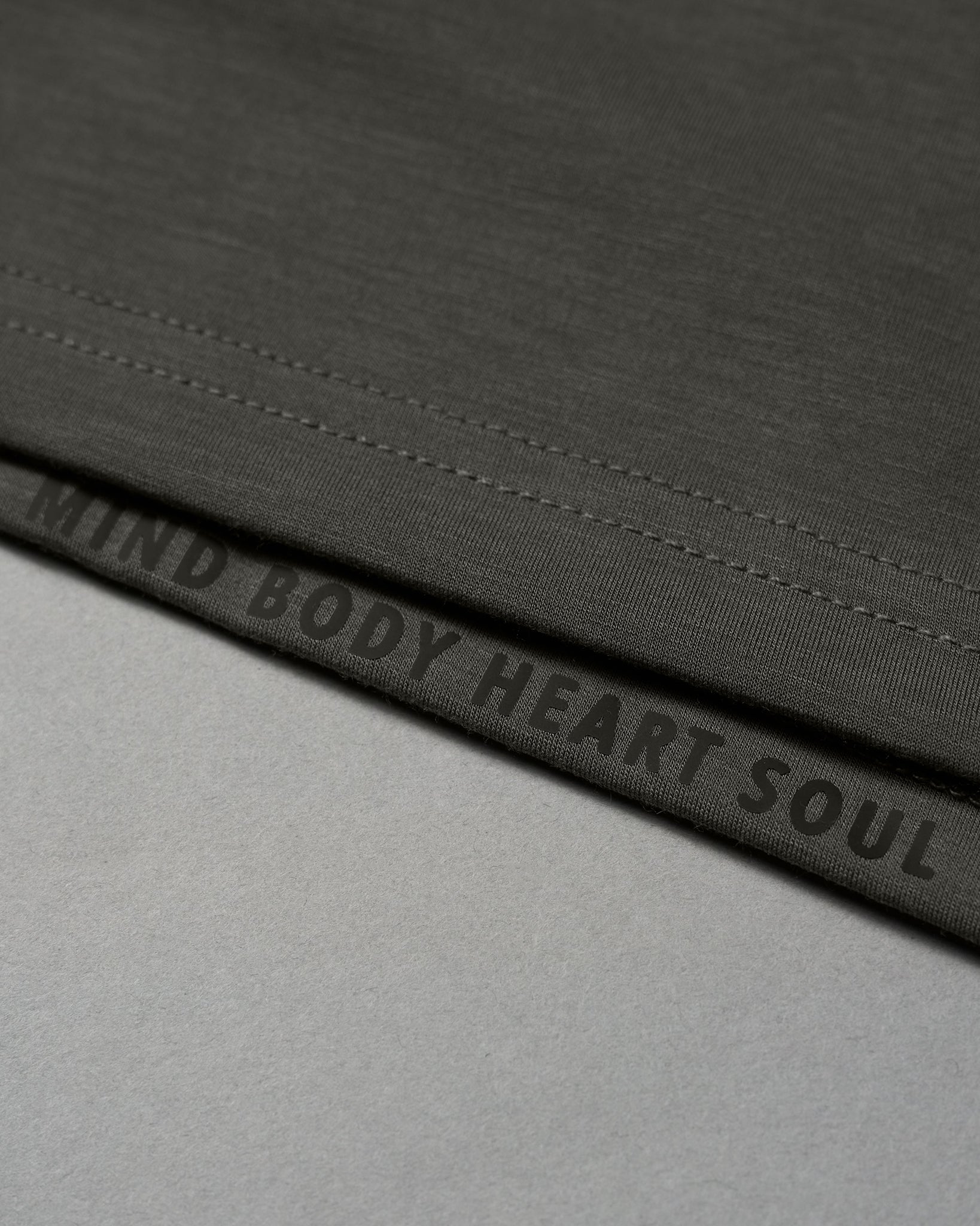 The hem of a dark green shirt prominently displays the phrase 'MIND BODY HEART SOUL' in black, set against a light gray background, conveying a holistic wellness theme.