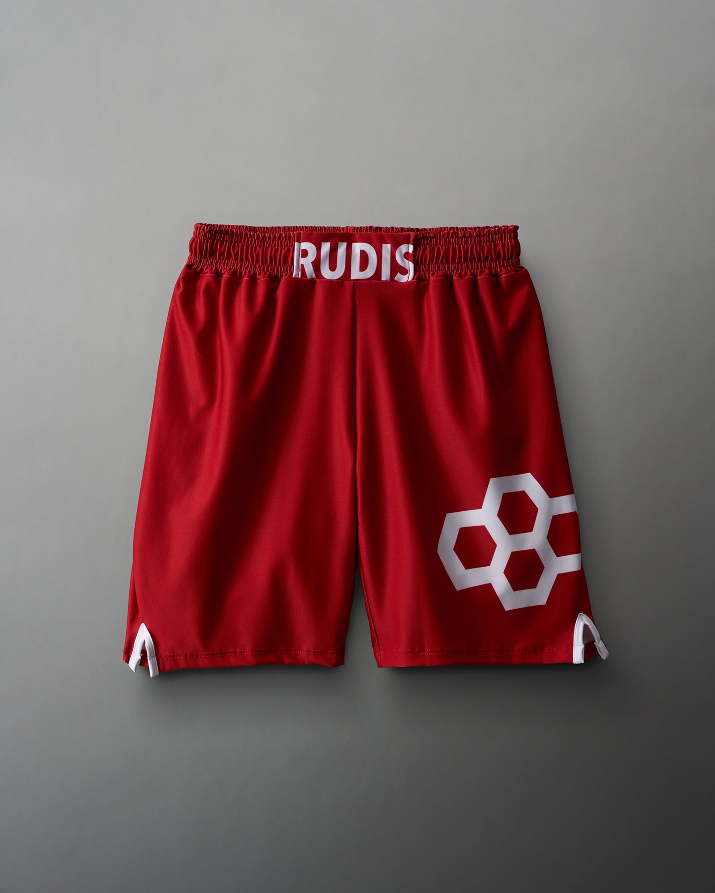 Red athletic shorts with a white 'RUDIS' logo on the waistband and interlocking hexagonal design on the side, set against a muted gray background.