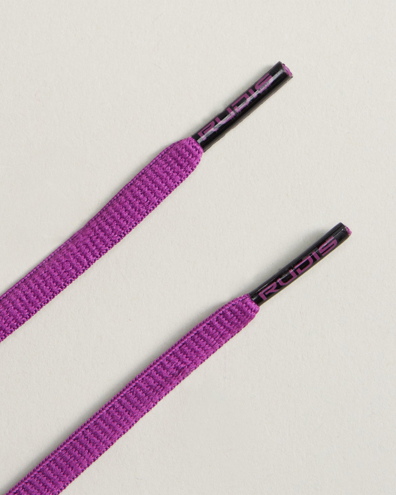 Two vibrant purple shoelaces featuring black aglets with the brand name RUDIS printed on them placed on a light-colored surface