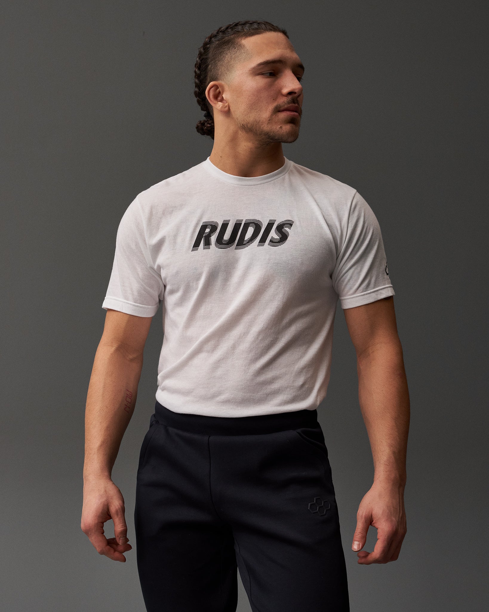 A confidently posed young man with braided hair wears a white 'RUDIS' t-shirt and black sweatpants, highlighted against a soft gray background.