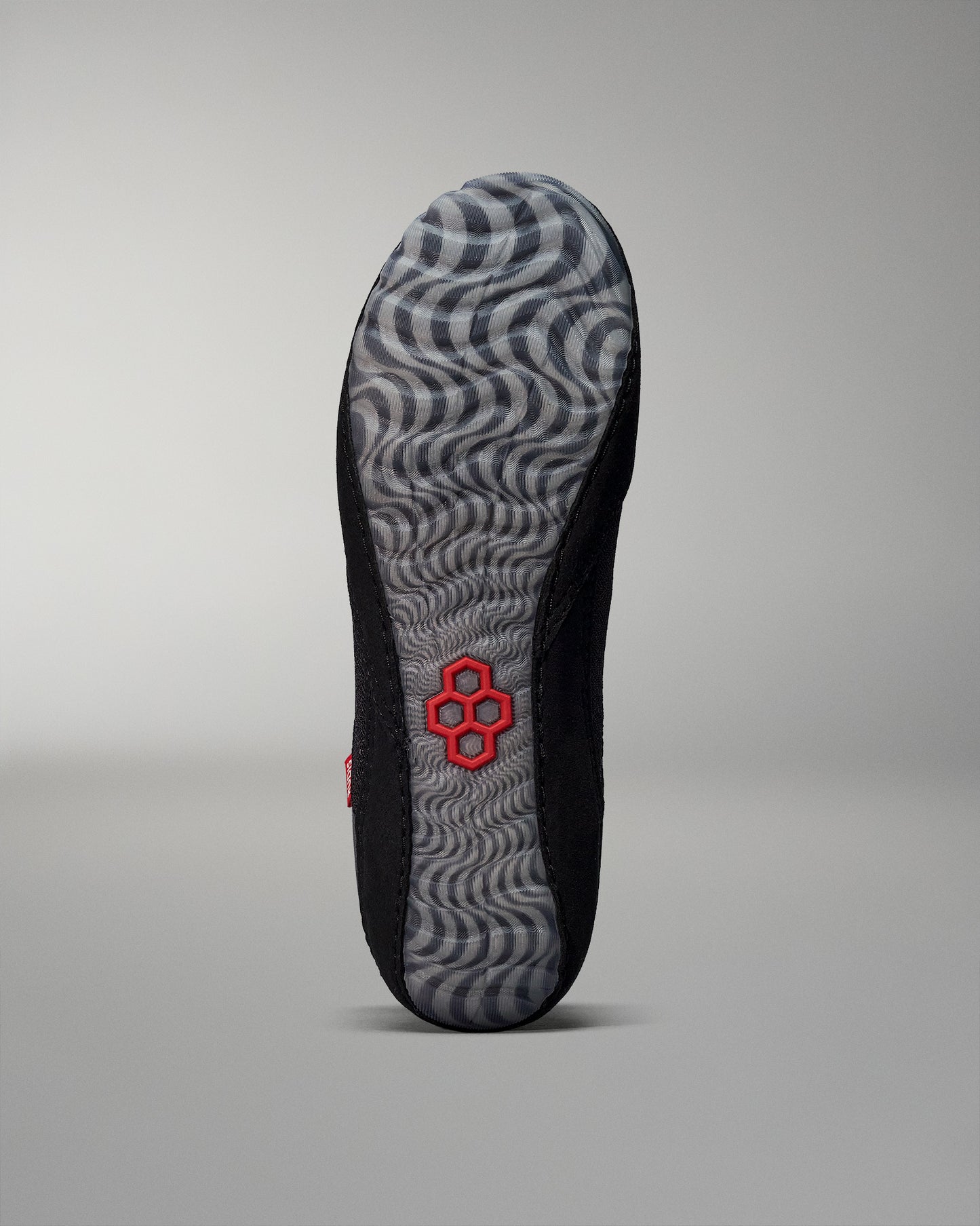 The sole of a shoe combines a textured black design with a wavy gray pattern and a central red logo, highlighting its performance-oriented features.