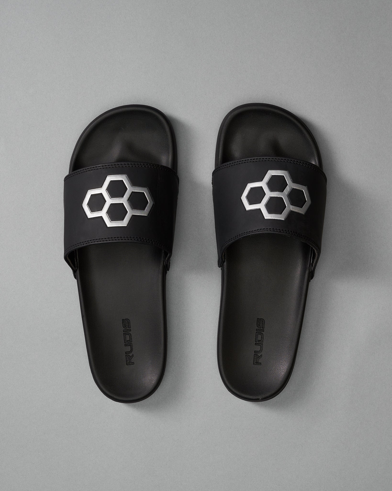 Black slide sandals with a honeycomb design and 'RUDIS' branding, showcased against a soft gray background.