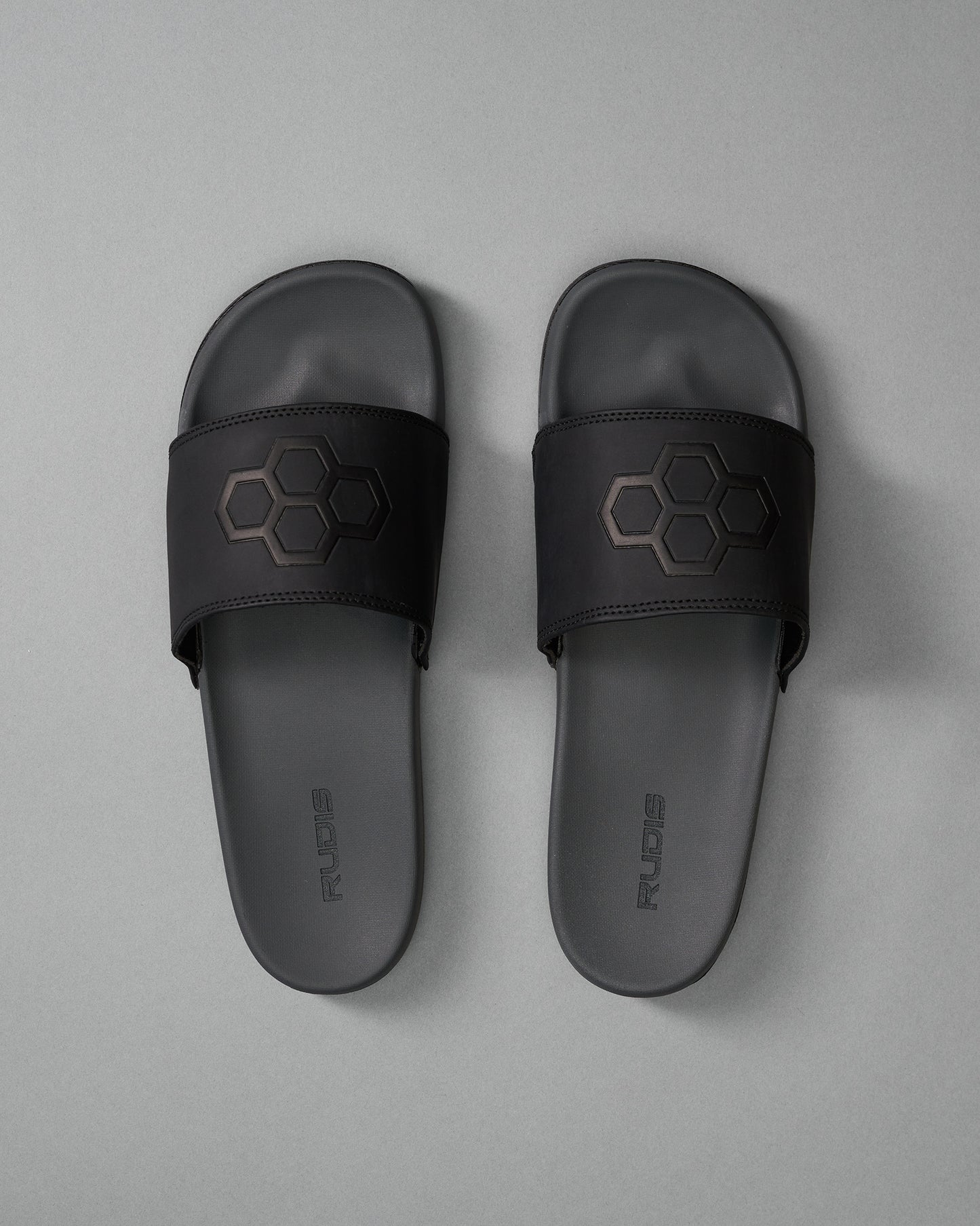 Black slide sandals showcasing a hexagonal logo on the strap, with a smooth footbed against a muted gray background.