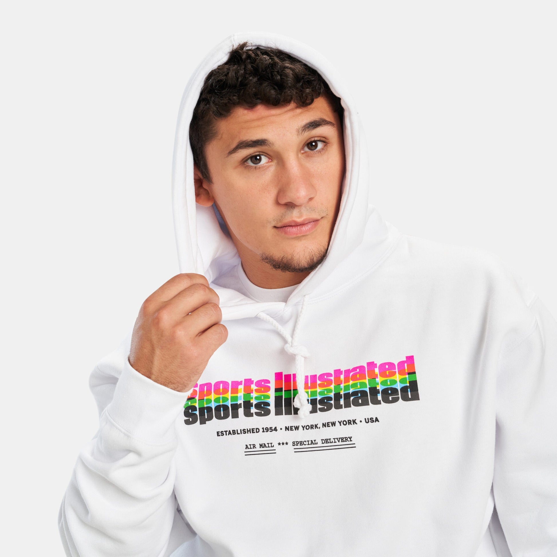 A young man in a stylish white hoodie featuring colorful Sports Illustrated branding posing thoughtfully