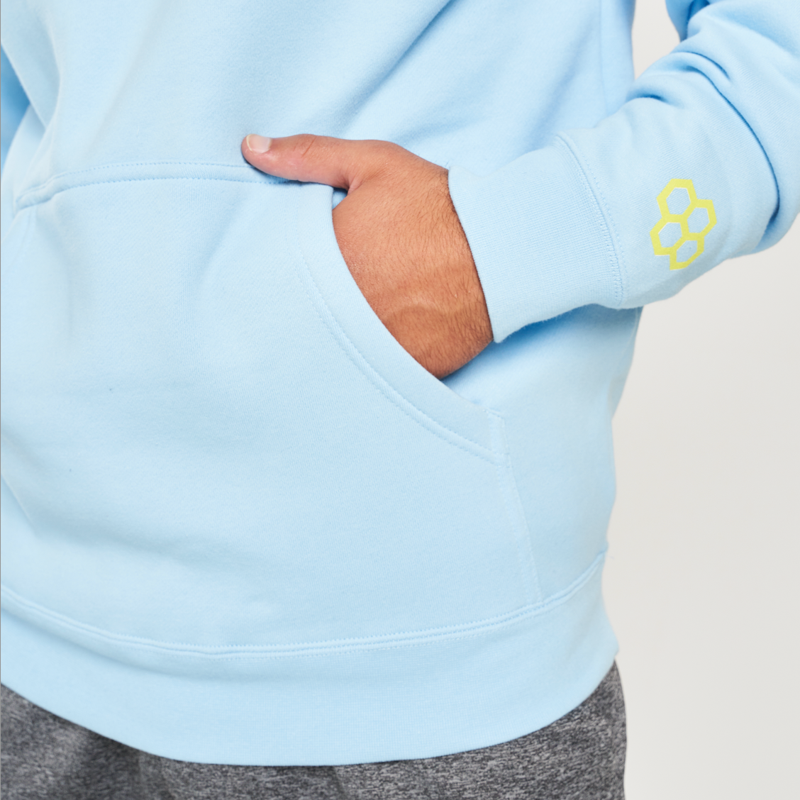 This image features a close-up of a persons hand resting in the pocket of a light blue hoodie accentuating a casual and comfortable style with a minimalistic design and a yellow logo on the sleeve