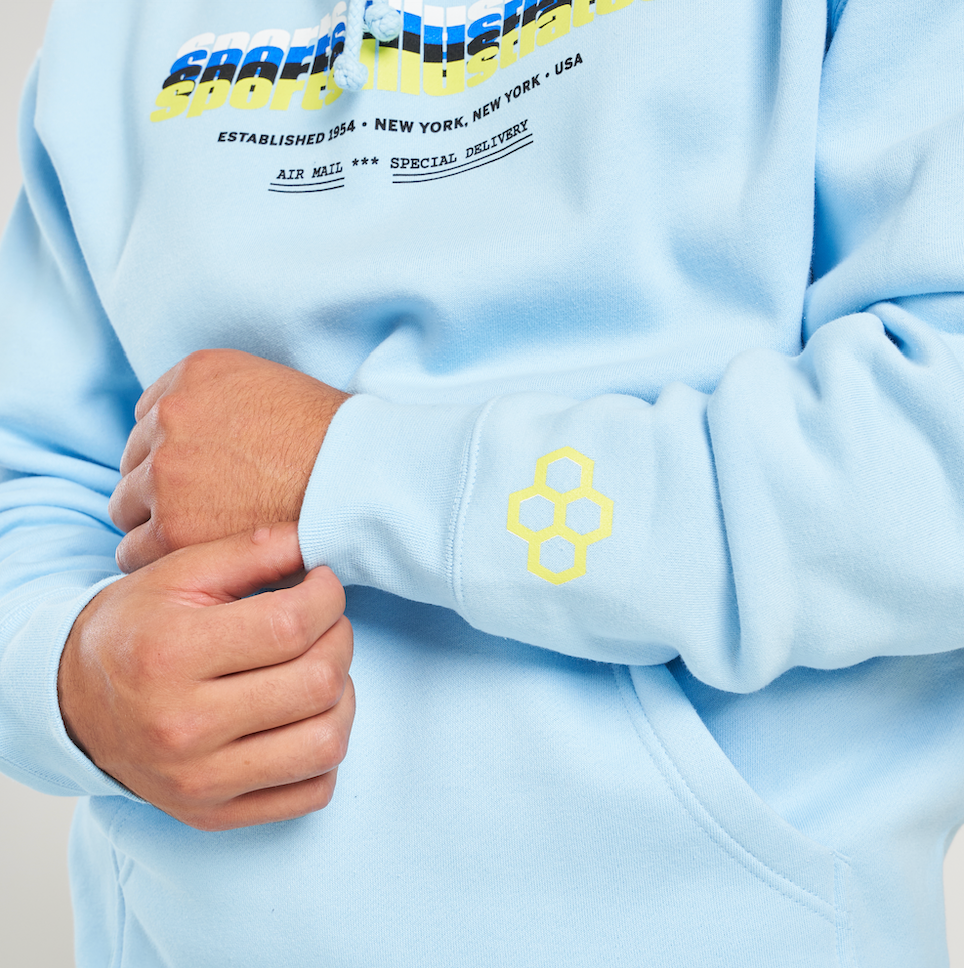 A close-up view of a person wearing a light blue hoodie with colorful graphic text and a yellow hexagonal logo on the sleeve