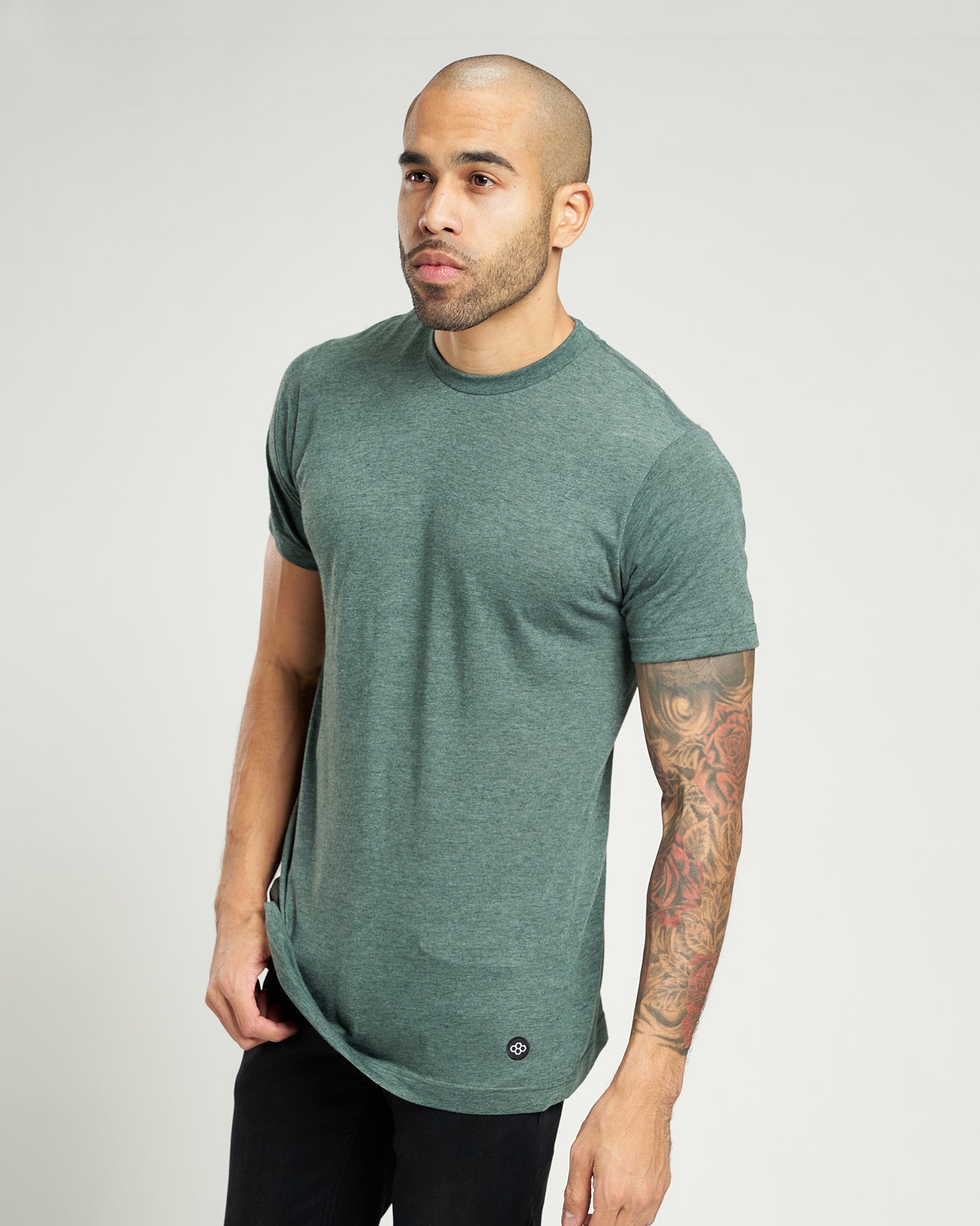 A man with a shaved head and a prominent beard is wearing a fitted green t-shirt showcasing his colorful tattoos on his arm against a neutral background