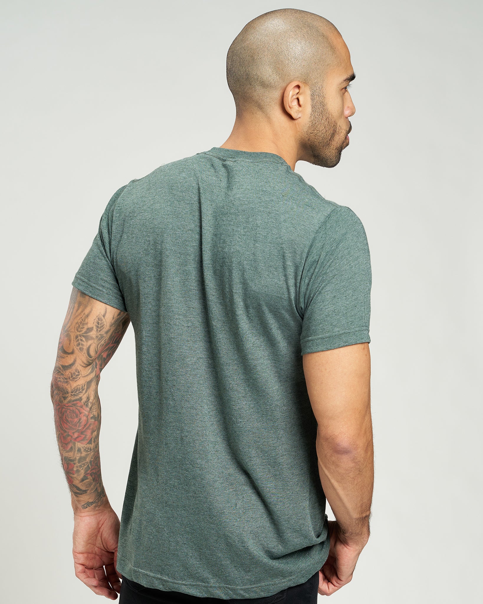 A man with a tattooed arm stands facing away wearing a fitted green shirt demonstrating a modern casual style