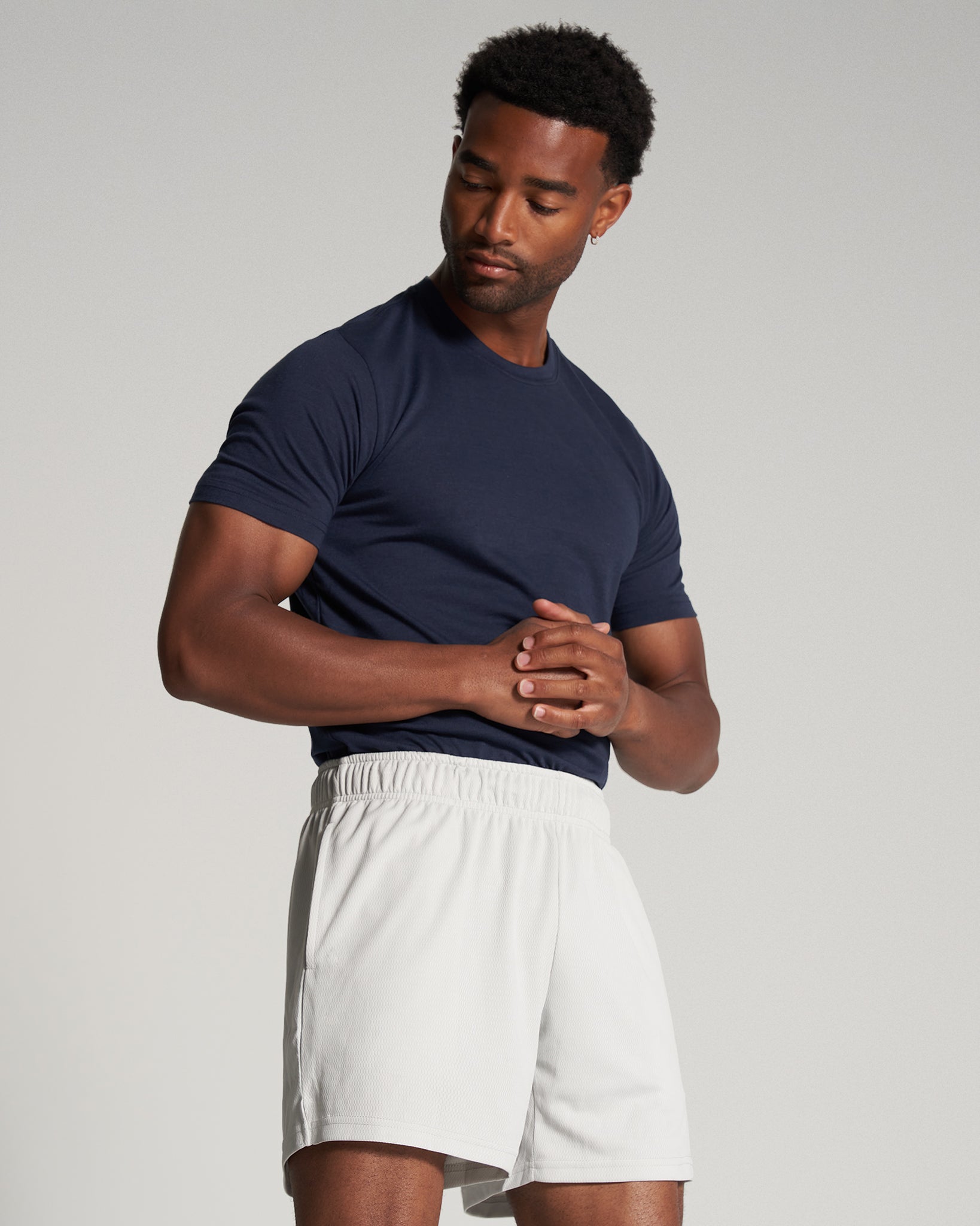 A man stands confidently with his hands clasped wearing a navy blue t-shirt and light beige shorts against a simple gray background