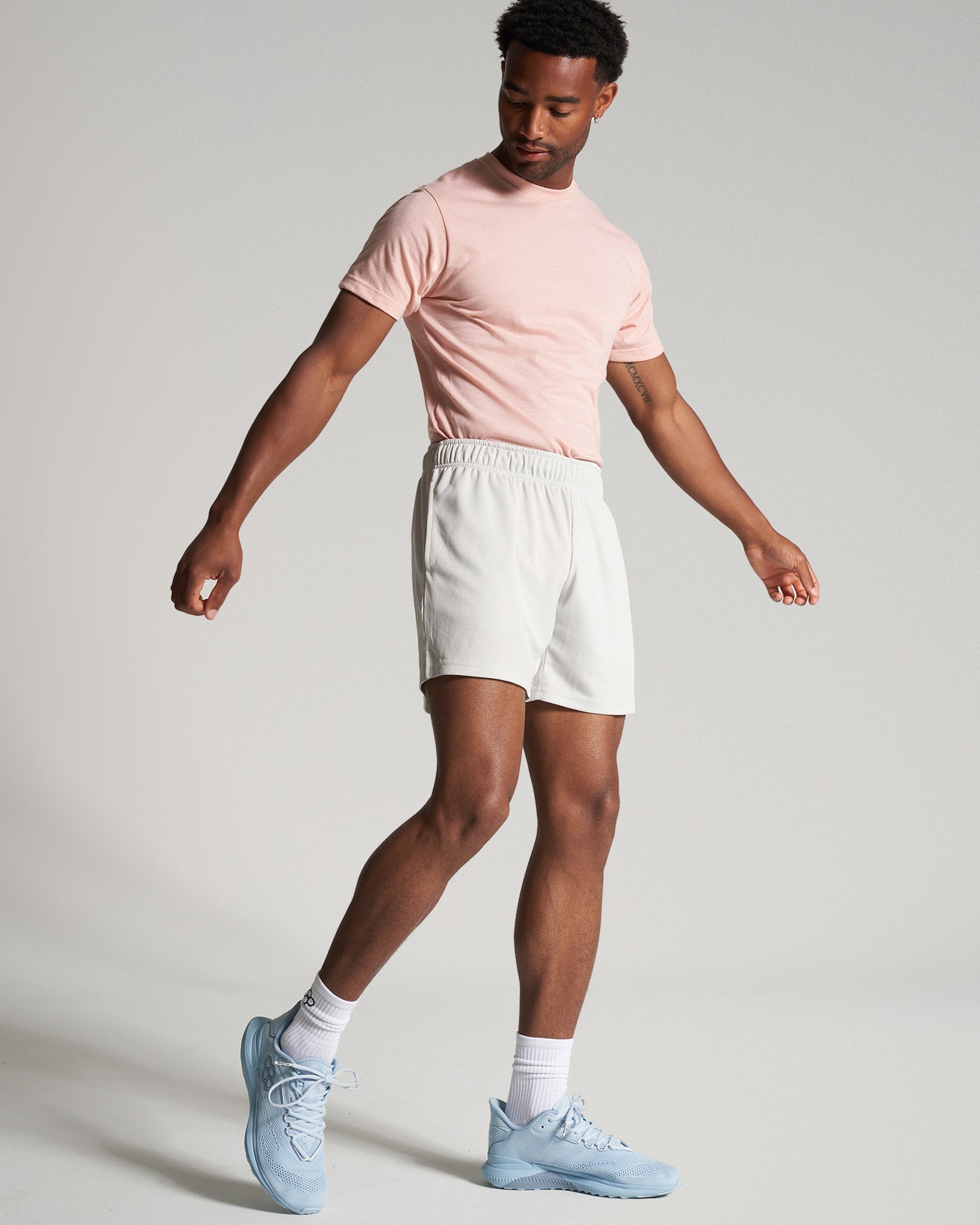 A man in a pink t-shirt and light shorts strikes a casual pose showcasing athletic footwear in a minimalist setting