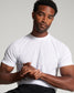 A muscular individual wearing a white T-shirt is seated displaying a casual yet confident demeanor with hands clasped together at the waist against a neutral background