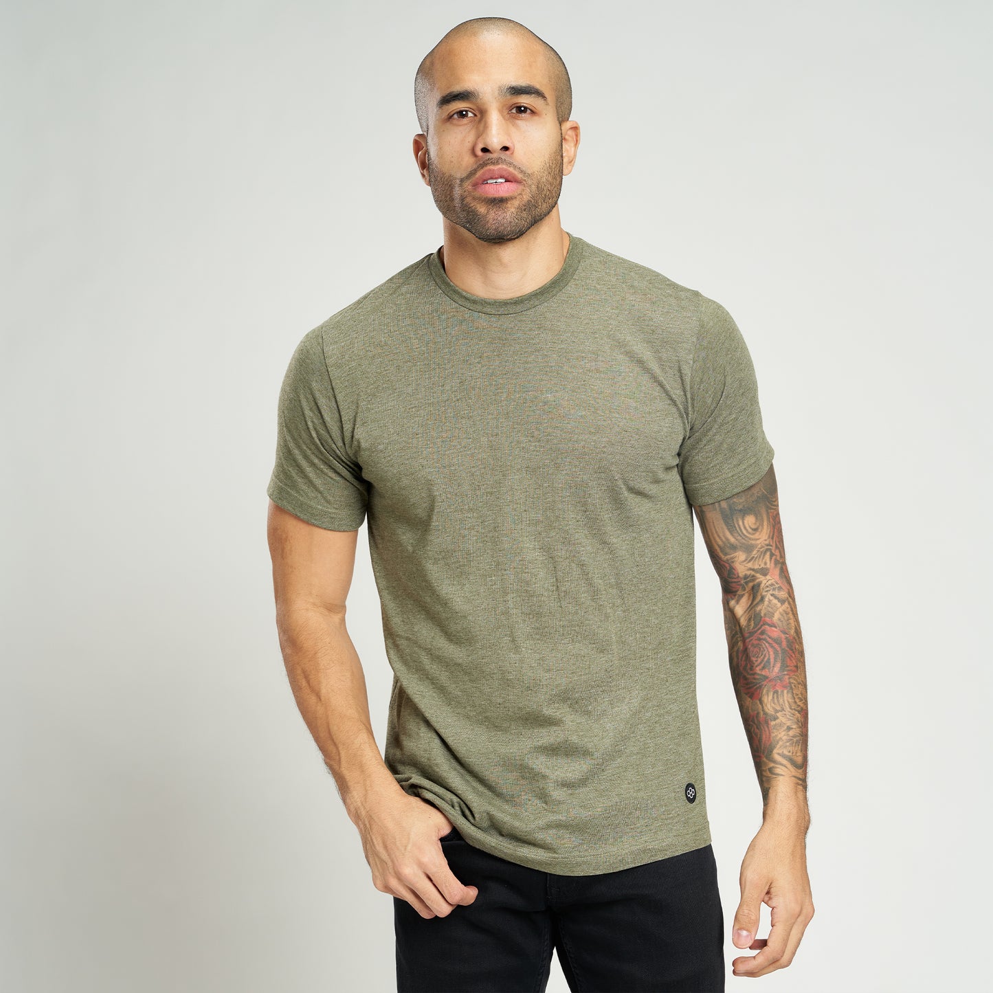 A man stands confidently in a fitted olive green t-shirt showcasing his tattoos and casual style against a light background