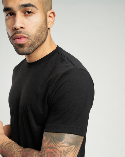 A close-up portrait of a man with a beard and tattoos wearing a fitted black t-shirt against a neutral background