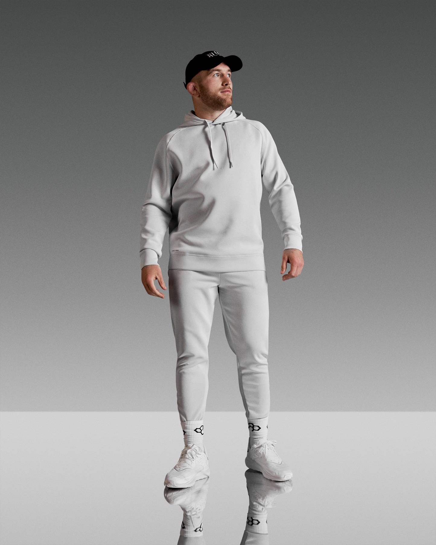 A man in a light gray hoodie and jogger pants stands confidently against a dark and light gray background, wearing white sneakers and a black cap.