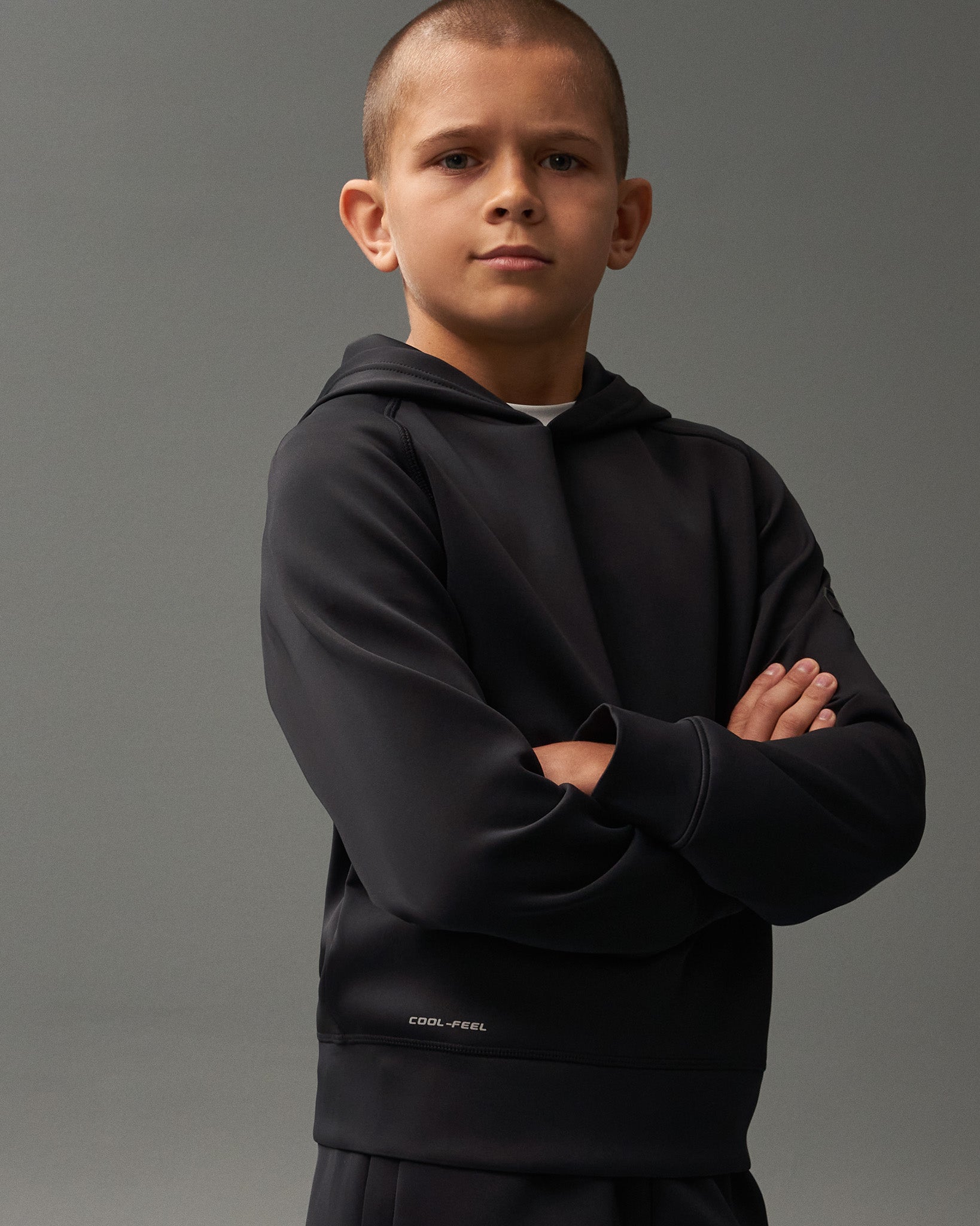 A young boy confidently poses with arms crossed in a black hoodie featuring 'COOL-FEEL' branding, set against a neutral gray background.