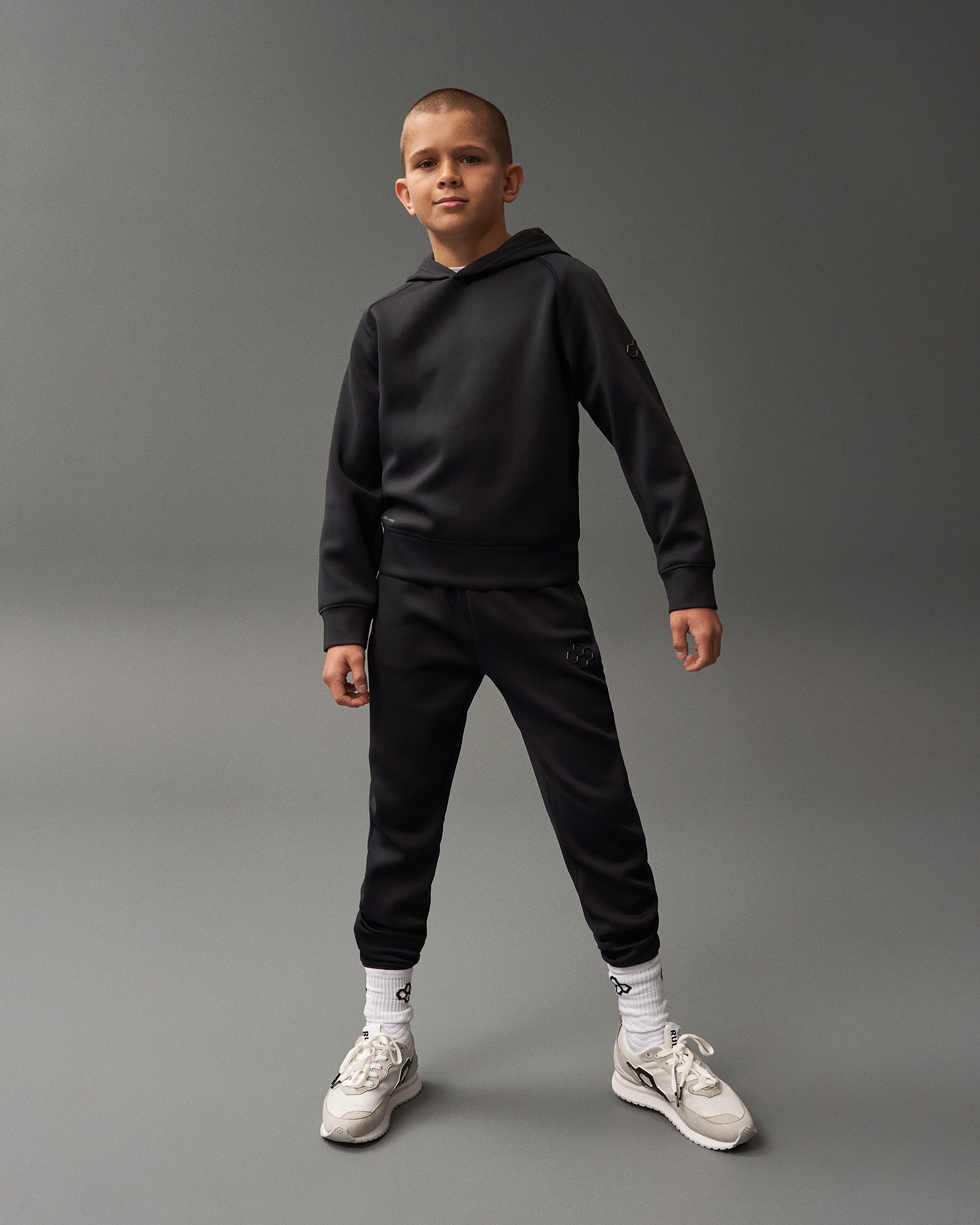 A young boy confidently poses in a black hoodie and pants with visible white socks and light sneakers against a muted gray background.