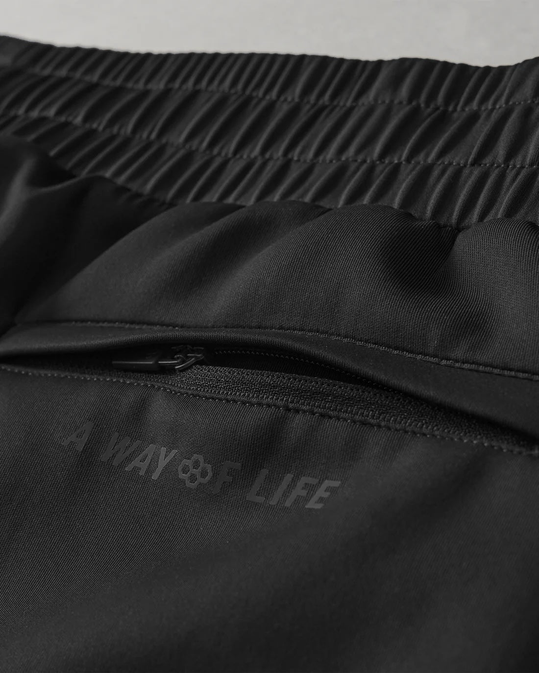 Black athletic shorts with an elastic waistband feature a zippered pocket and the phrase 'A WAY OF LIFE', detailed against a neutral gray background for emphasis.