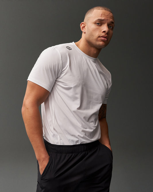 A confident man in a fitted white t-shirt with a logo and black athletic shorts stands against a dark gray background, showcasing his athletic build and a unique scar on his forehead.