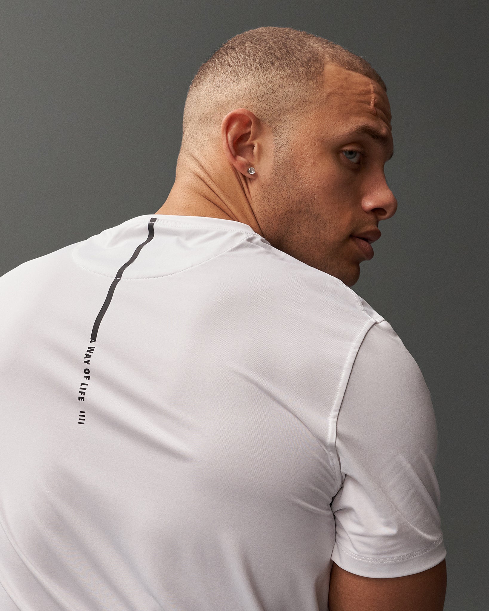 A man poses in a white athletic shirt adorned with vertical black text on the back against a gray background, highlighting his profile and modern style.