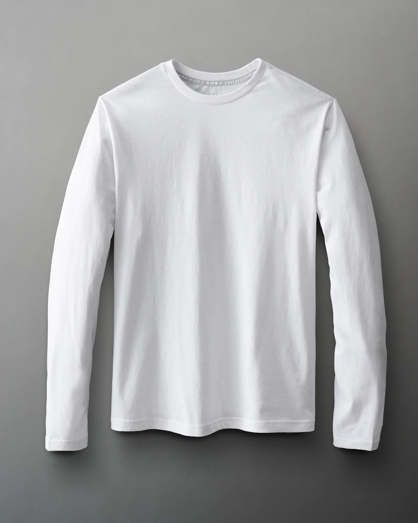 A plain white long-sleeve shirt with a classic crew neckline and slightly curved hem is displayed against a subtle gray background, highlighting its simple design and soft texture.