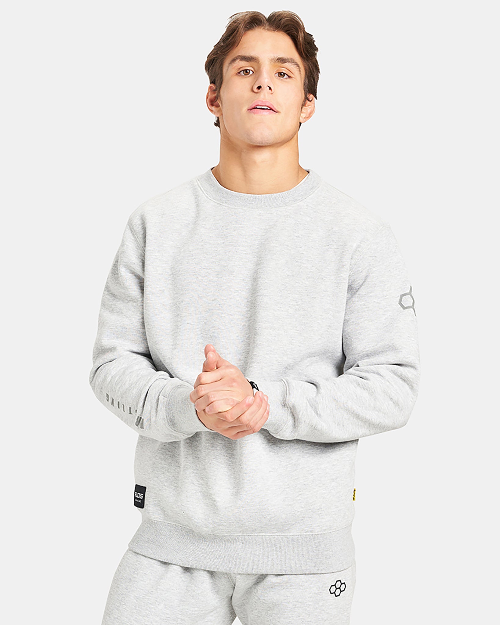 A young man wearing a light gray sweatshirt stands confidently with his hands clasped showcasing a casual stylish look suitable for everyday wear