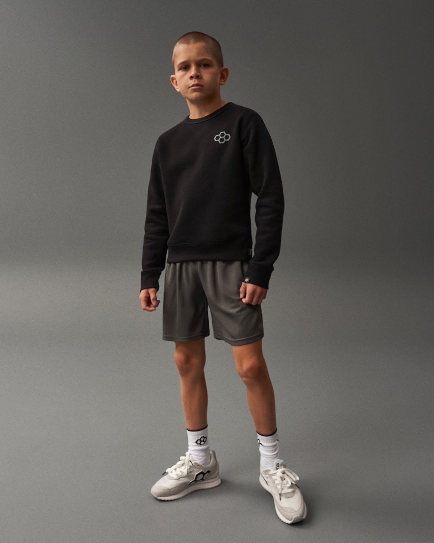 A young boy stands confidently in a black sweatshirt and gray shorts wearing white sneakers and patterned socks against a minimalist gray background