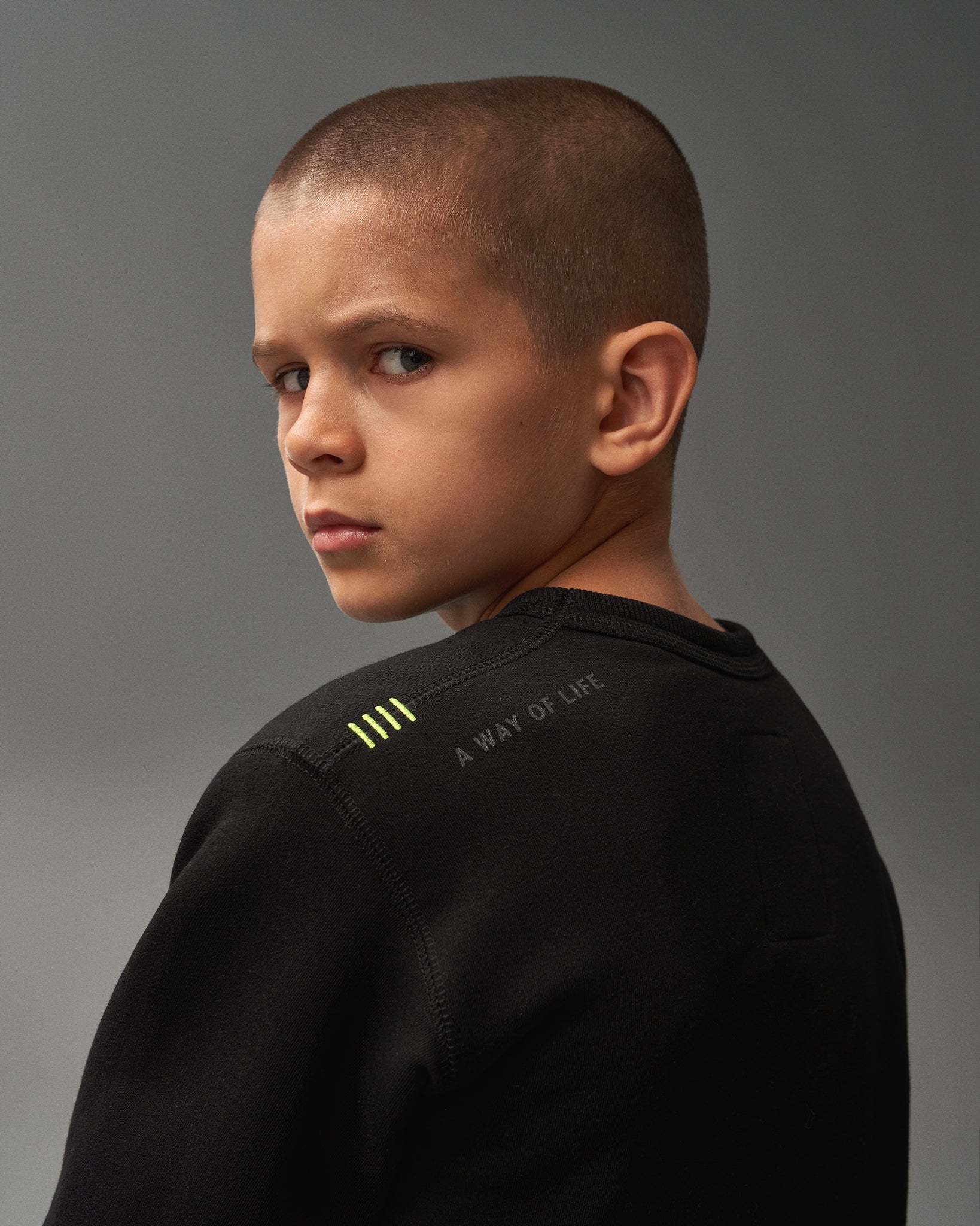 A boy with a buzz cut gazes intently over his shoulder wearing a black sweatshirt with a distinctive phrase and design elements