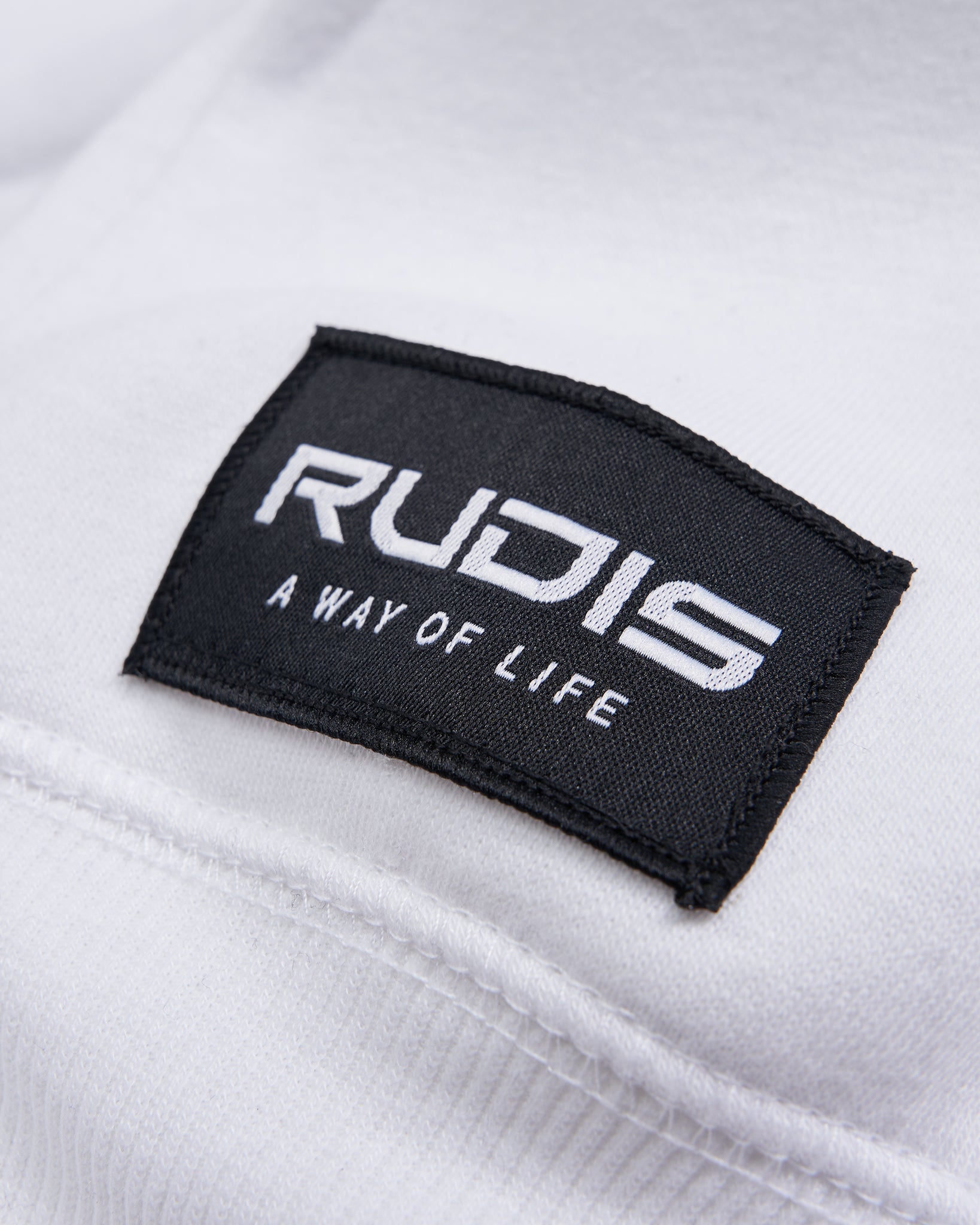 Close-up of a black tag sewn onto a white garment featuring the word RUDIS and the phrase A WAY OF LIFE