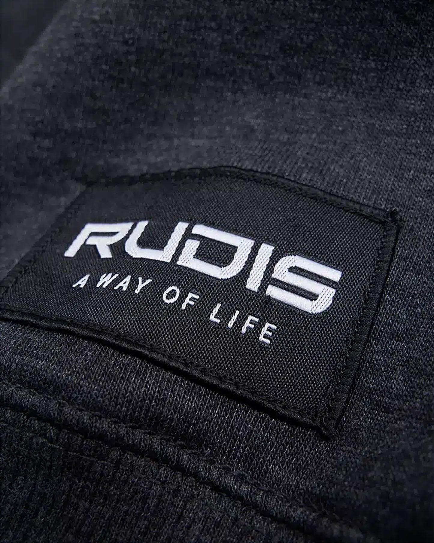 A close-up of a black clothing label featuring the brand name RUDIS and the phrase A WAY OF LIFE in white text set against a textured fabric background