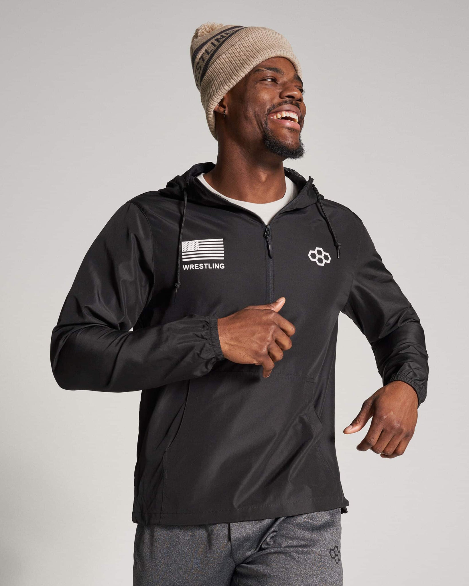 A smiling man wearing a black athletic jacket and a beige beanie engaged in a running motion against a light gray background